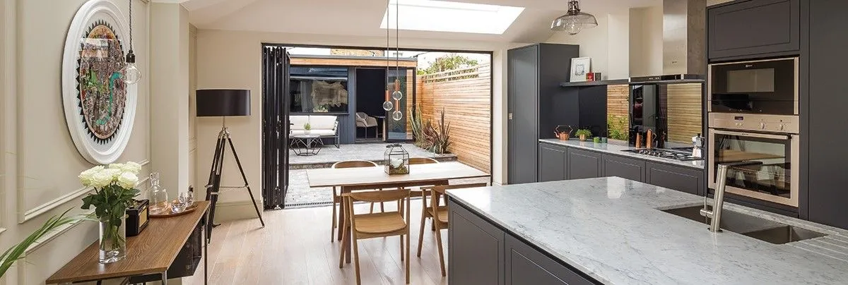 A modern, open-plan kitchen and dining area in a renovated Victorian house, featuring a marble island, dark cabinetry, and bi-fold doors opening onto a patio.